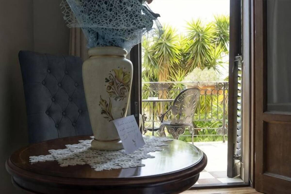 A table with a vase, lace, and a card, a chair, and a view of a balcony with plants.