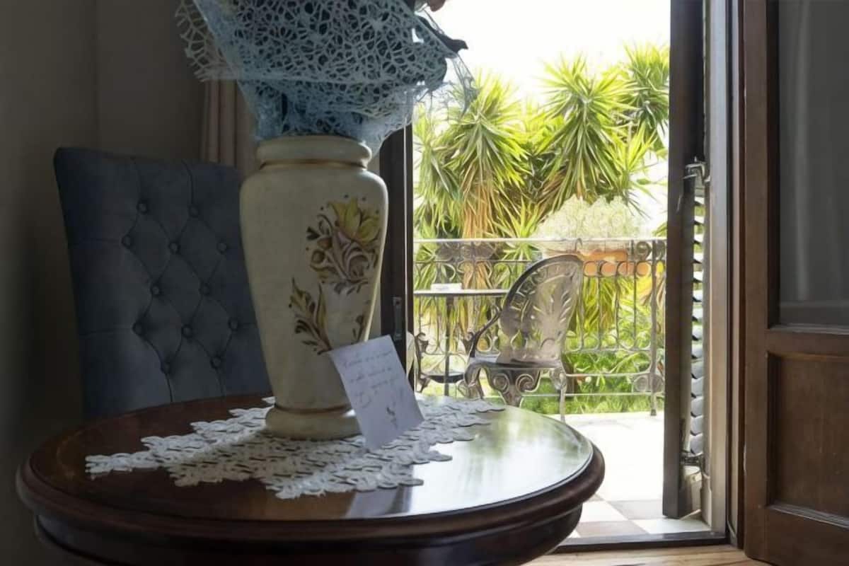 A table with a vase, lace, and a card, a chair, and a view of a balcony with plants.