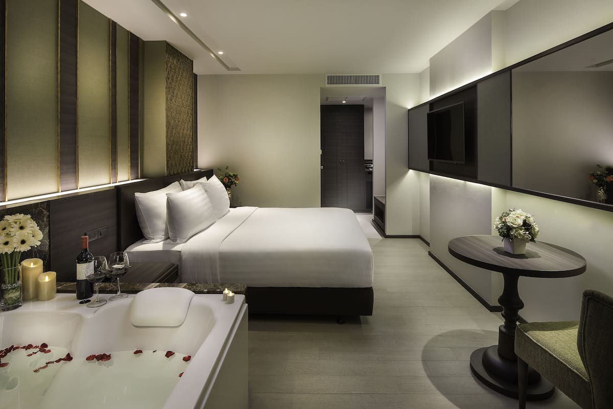 A modern hotel room with a large bed, a sitting area with a round table, a bathtub, and a TV mounted on the wall.