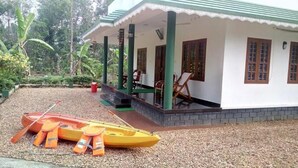 A white building with green columns and a covered porch. There are two boats and life jackets on the ground. - Kuttickattil Gardens Home Stay (Kottayam)