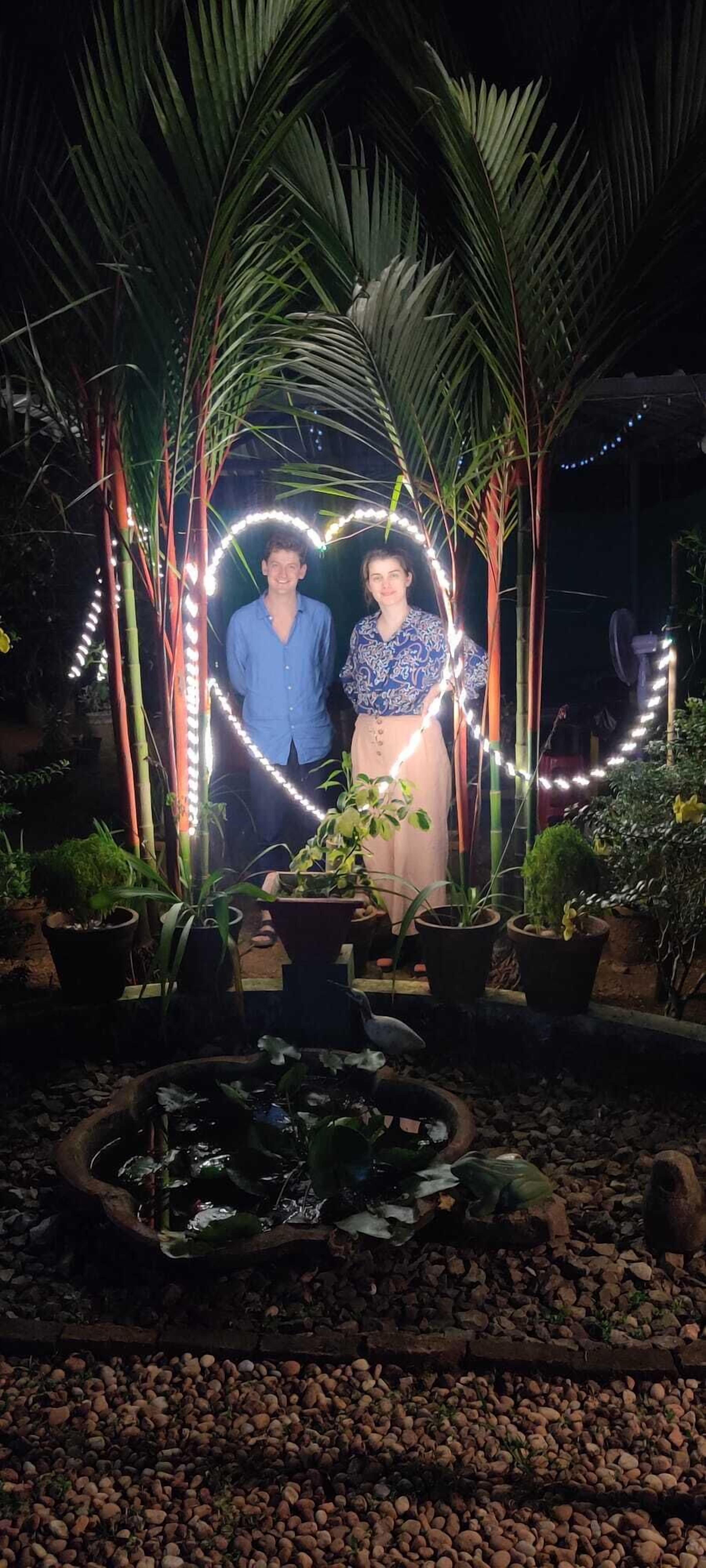 A couple standing under a canopy of palm trees with string lights forming a heart shape.