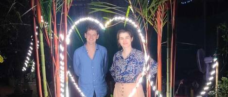 A couple standing under a canopy of palm trees with string lights forming a heart shape.