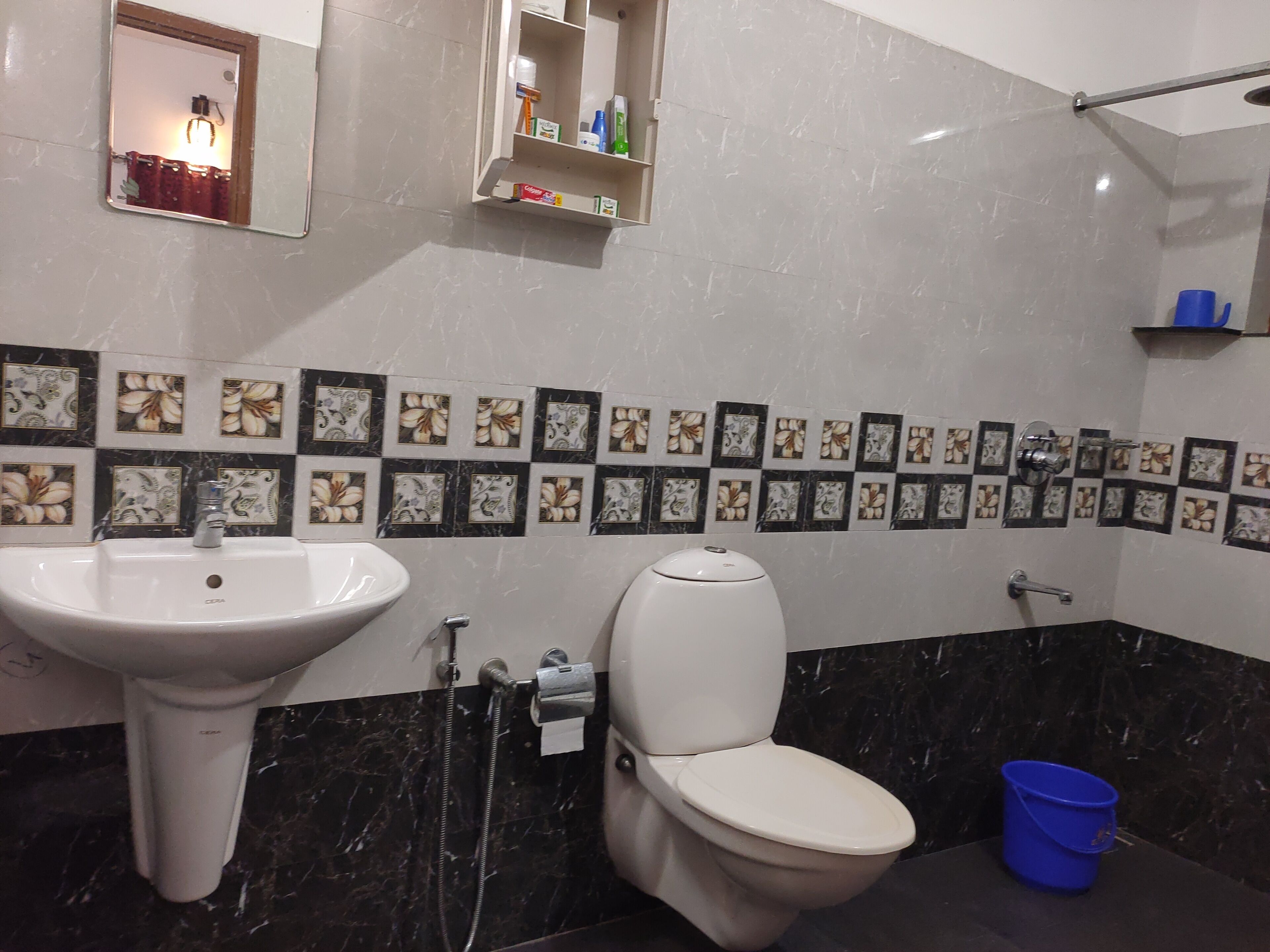 A bathroom with a white sink, toilet, and shower. There are decorative tiles on the wall and a blue bucket on the floor.
