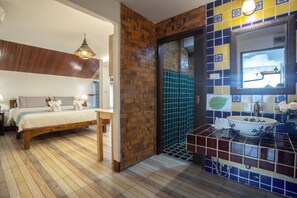 Une chambre à coucher avec un lit en bois, une table de chevet et une commode. La salle de bain est équipée d’un lavabo, d’un miroir et de murs carrelés.