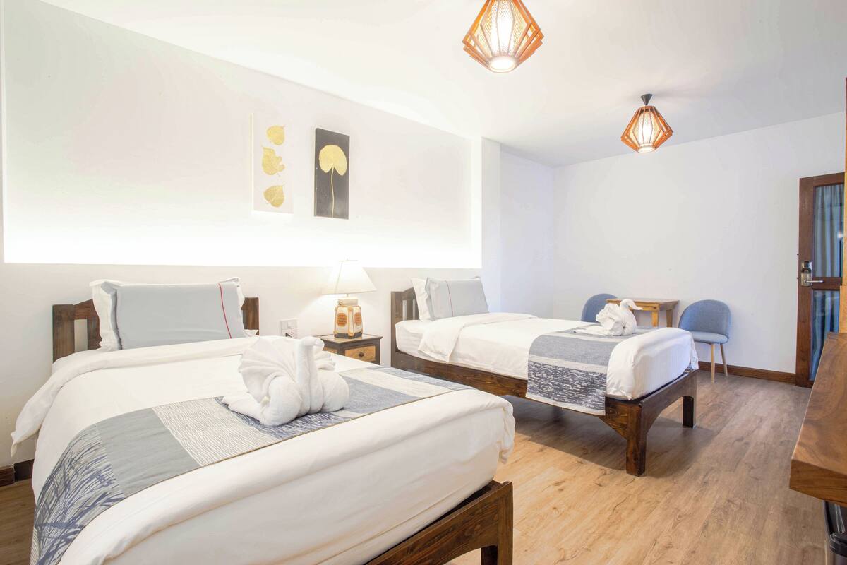 Ein Hotelzimmer mit zwei Betten, Holz-Kopfteilen, einem kleinen Tisch, einem Sessel und einer Wandlampe.