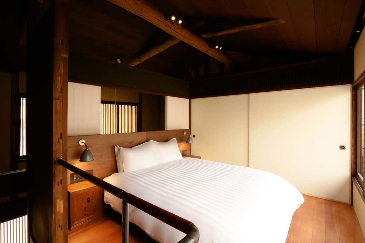Een slaapkamer met een houten hoofdeinde, een bed met witte beddengoed, een nachtkastje met een lamp en een raam met verticale jaloezieën.