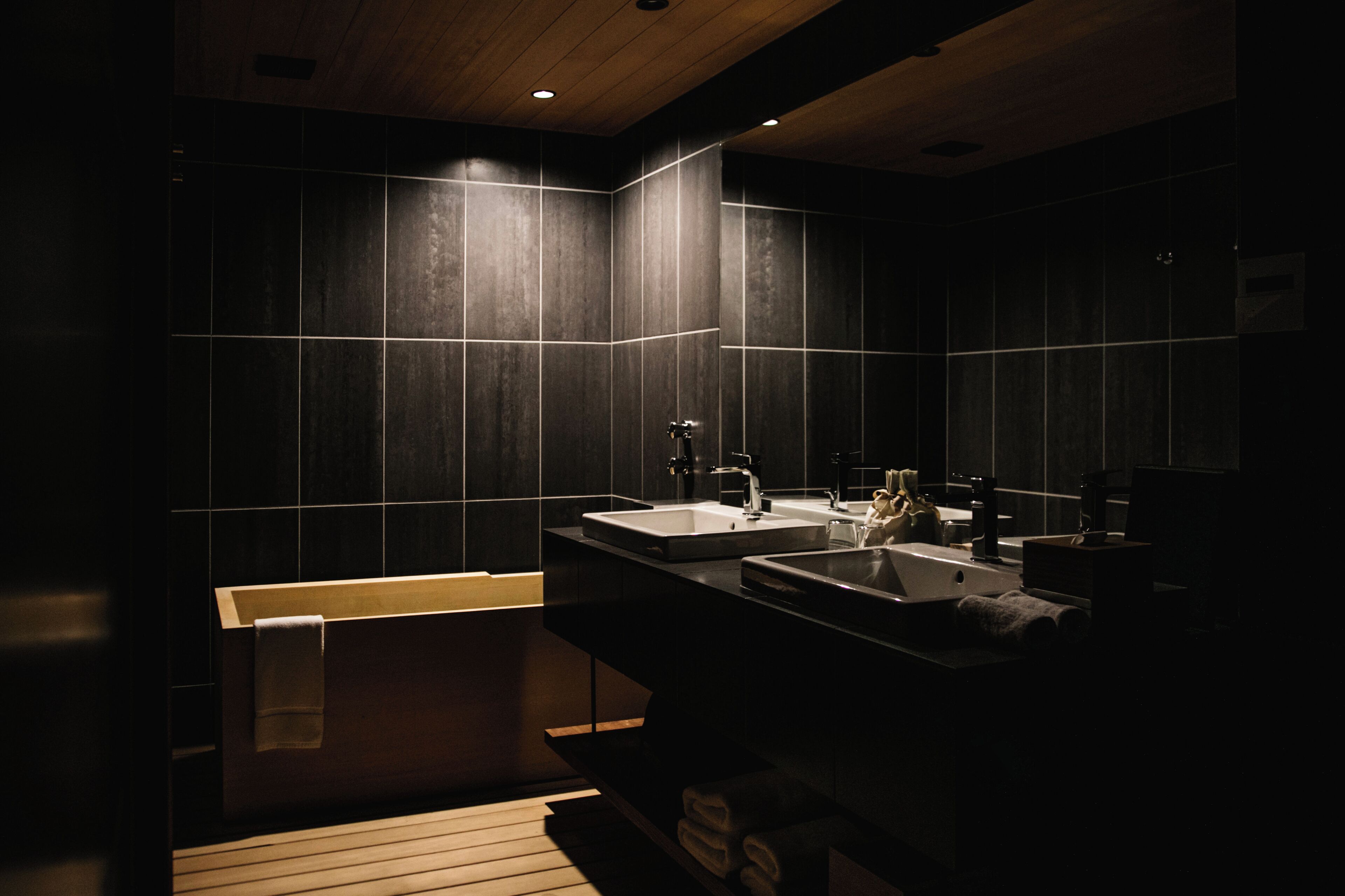 A modern bathroom with dark tiles, two sinks, and a wooden bench.