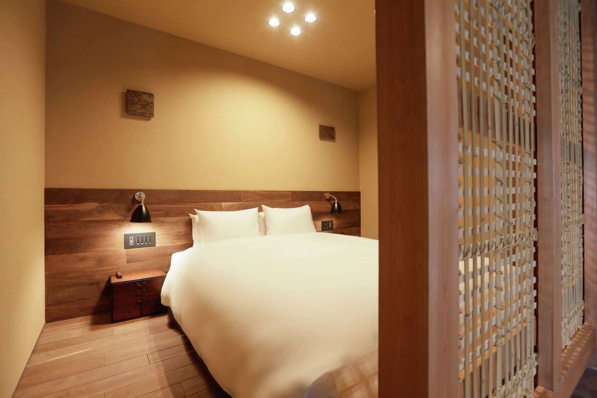 Een hotelkamer met een houten hoofdbord, een bed met witte lakens, twee bedlampjes en een klein nachtkastje met een lamp.
