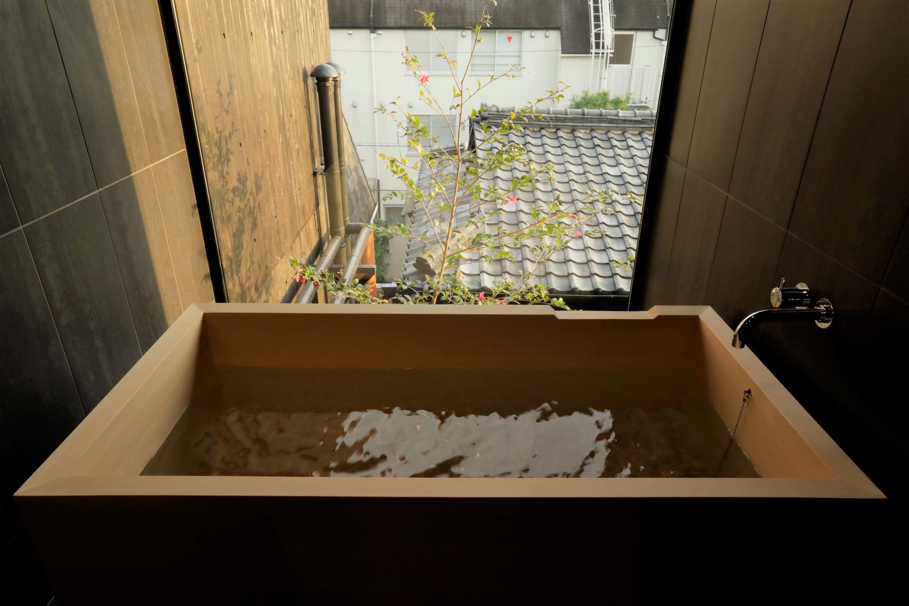 A wooden bathtub with a view of a tiled roof and a plant outside.