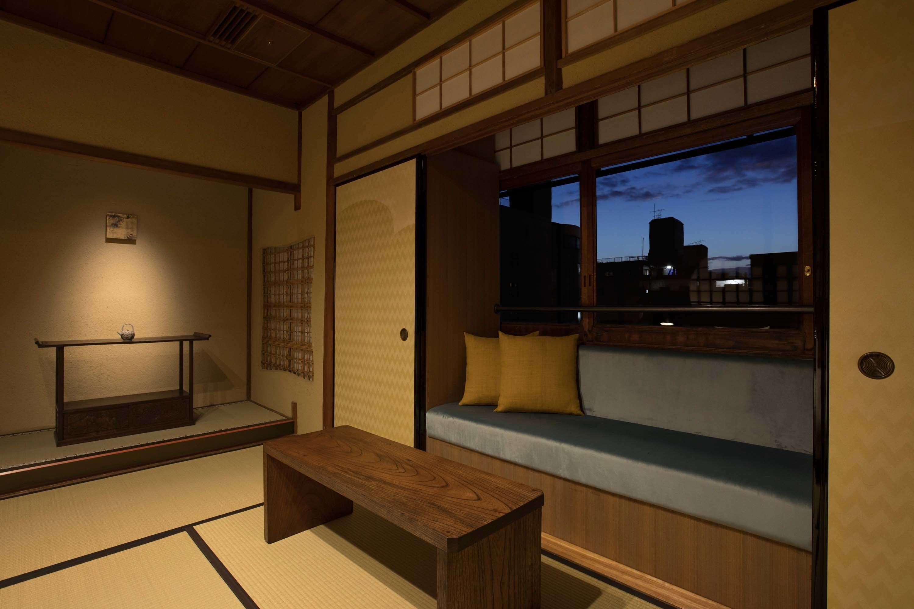 Une pièce de style japonais traditionnel, avec un sol recouvert de tatamis, une basse table en bois, un coin salon agrémenté de coussins, et une vue sur le paysage urbain au crépuscule.