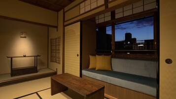 Une pièce de style japonais traditionnel, avec un sol recouvert de tatamis, une basse table en bois, un coin salon agrémenté de coussins, et une vue sur le paysage urbain au crépuscule.