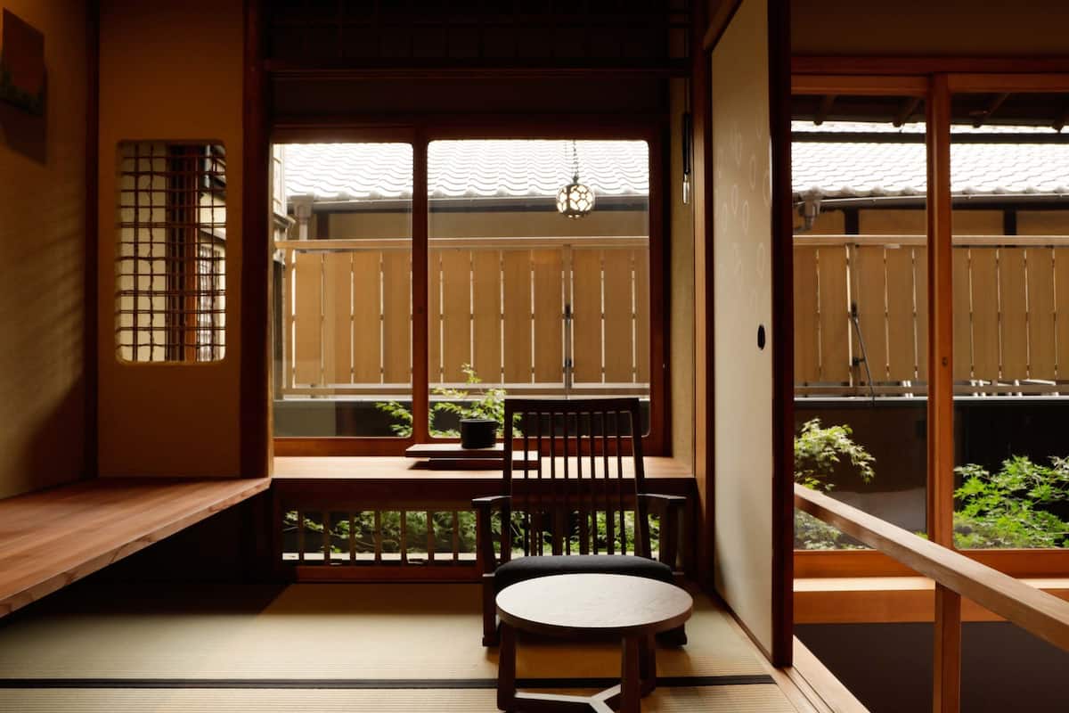Een traditionele Japanse kamer met tatami vloeren, een lage tafel, een stoel en een schuifdeur naar een balkon met uitzicht op groen.