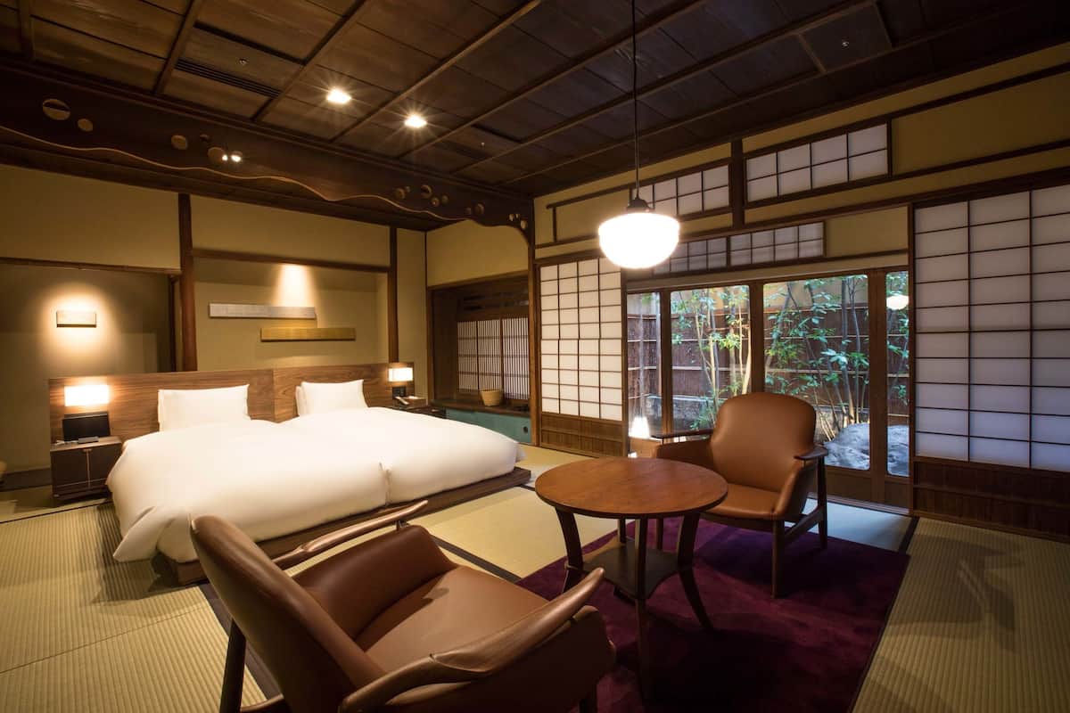 Een traditionele Japanse hotelkamer met tatami vloeren, een groot bed, een zithoek met een tafel en stoelen, en uitzicht op groen via de schuifdeuren.