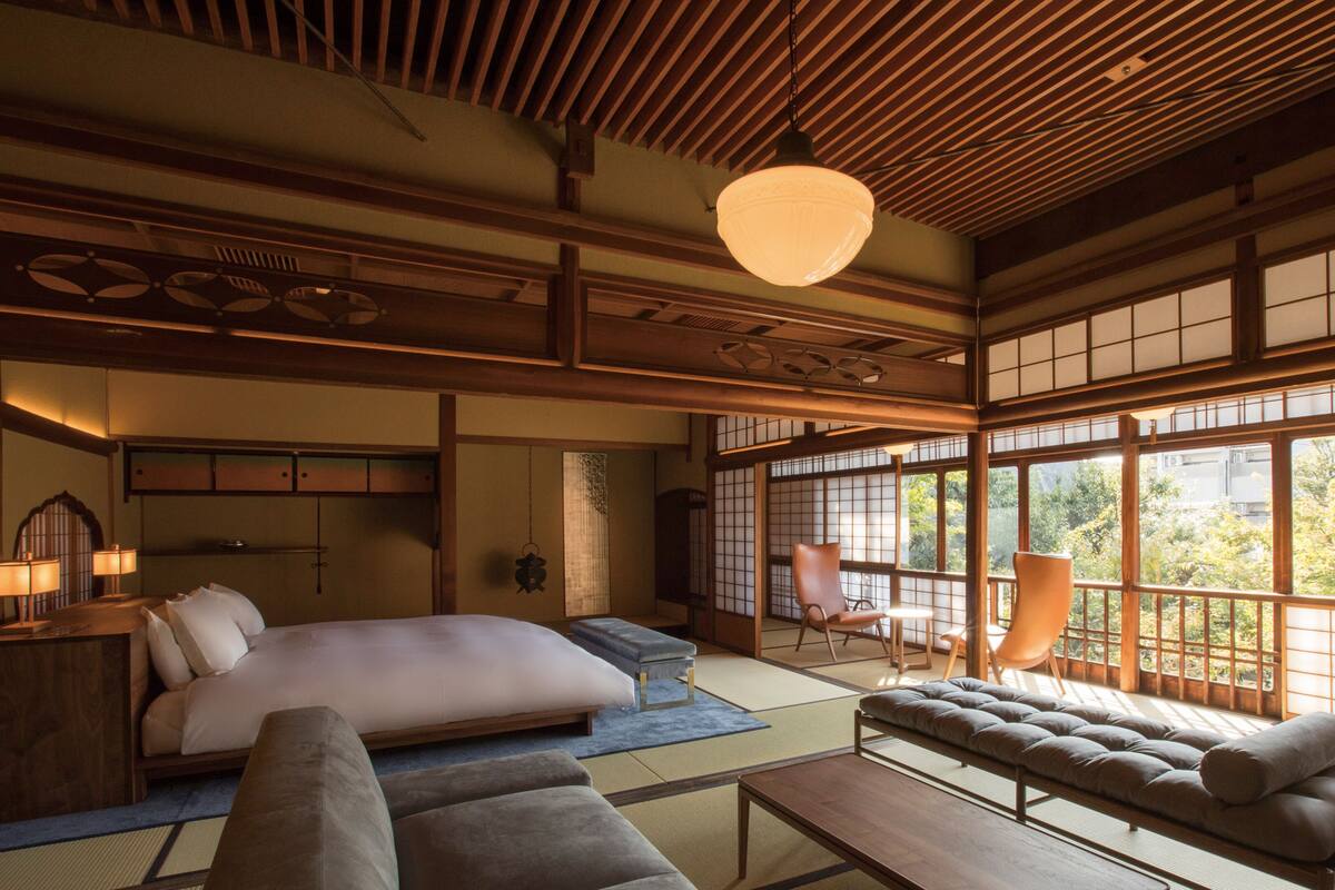 Een traditionele Japanse kamer met tatami vloerbedekking, een laag houten bed, een sofa en uitzicht op groen door grote shoji schermen.