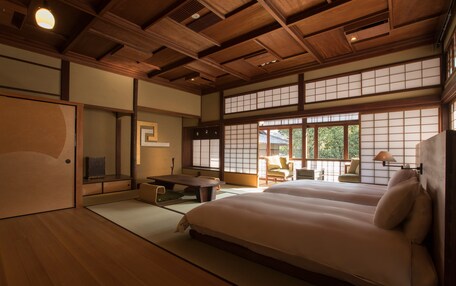 A traditional Japanese-style room with tatami flooring, a low wooden ceiling, and sliding shoji doors. There is a bed with white bedding, a small table, and a chair.