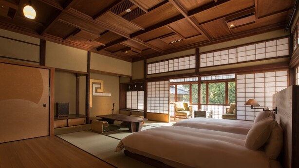 Una habitación tradicional japonesa con suelo de tatami, techo bajo de madera y puertas correderas shoji. Se observa una cama con ropa blanca, una pequeña mesa y una silla.