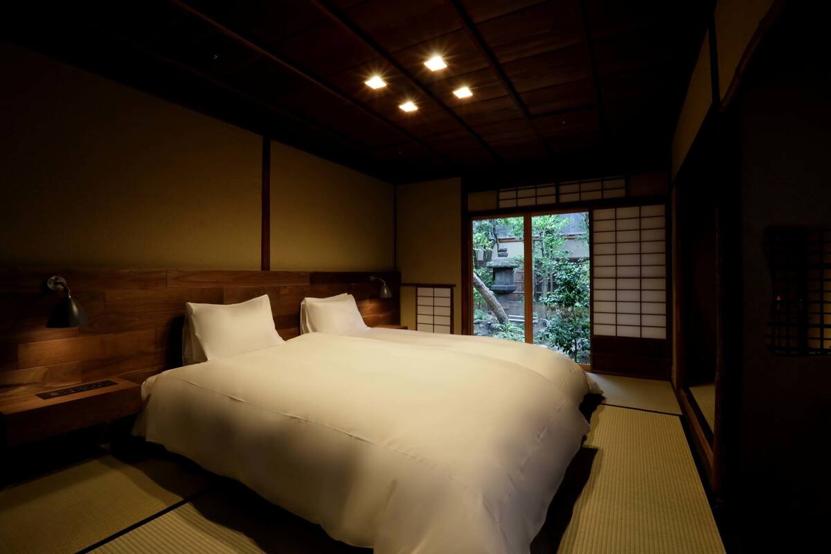 Een traditionele Japanse slaapkamer met tatami vloerbedekking, een houten hoofdbord en uitzicht op groen door een shoji scherm.