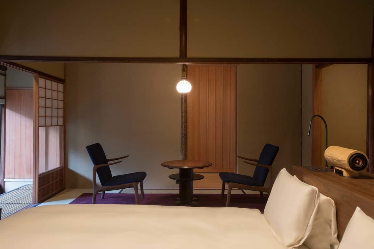 Een hotelkamer met een bed, twee stoelen, een klein rond tafeltje, een houten bureau en een traditionele Japanse schuifdeur.