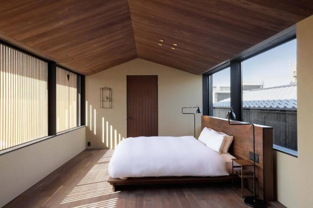 Een slaapkamer met een houten plafond, een bed, een nachtkastje, een lamp en grote ramen.