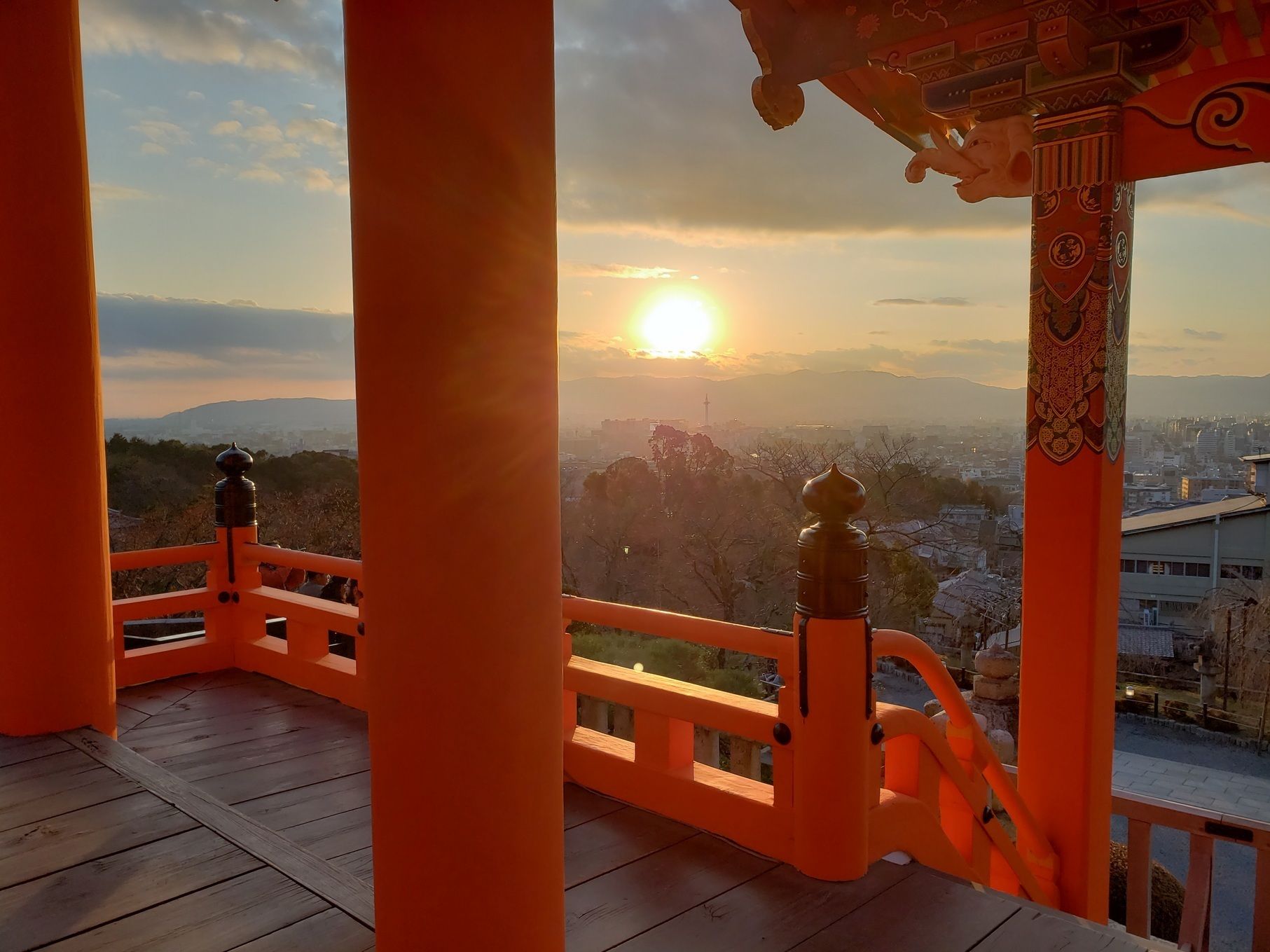 A wooden balcony with orange railings and a view of a cityscape during sunset.