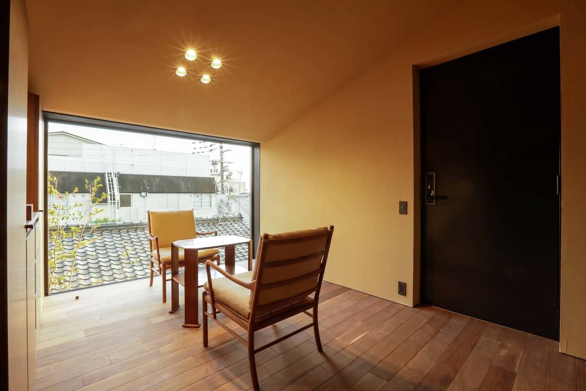 Een kamer met een houten vloer, een tafel met stoelen, een groot raam en een donkere deur.