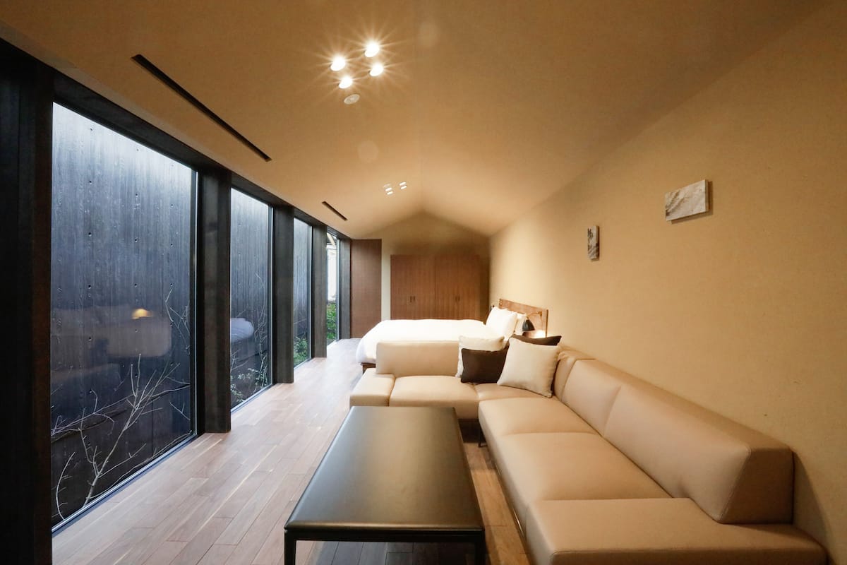 Een moderne slaapkamer met een groot bed, een bank en een tafel. Er zijn grote ramen die natuurlijk licht en uitzicht op de omgeving bieden.