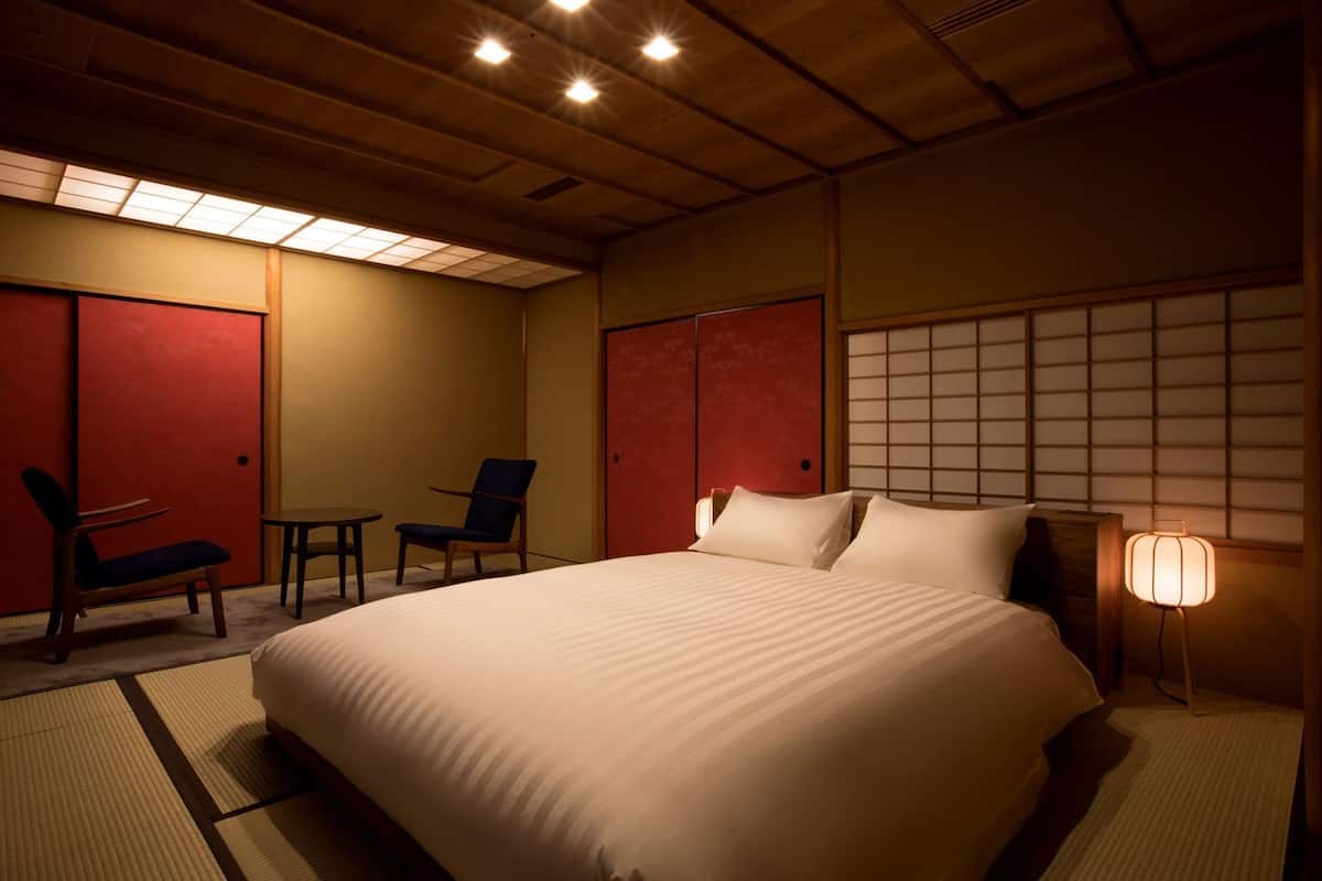 Een hotelkamer met een groot bed, twee stoelen, een tafeltje en een traditionele Japanse wand.