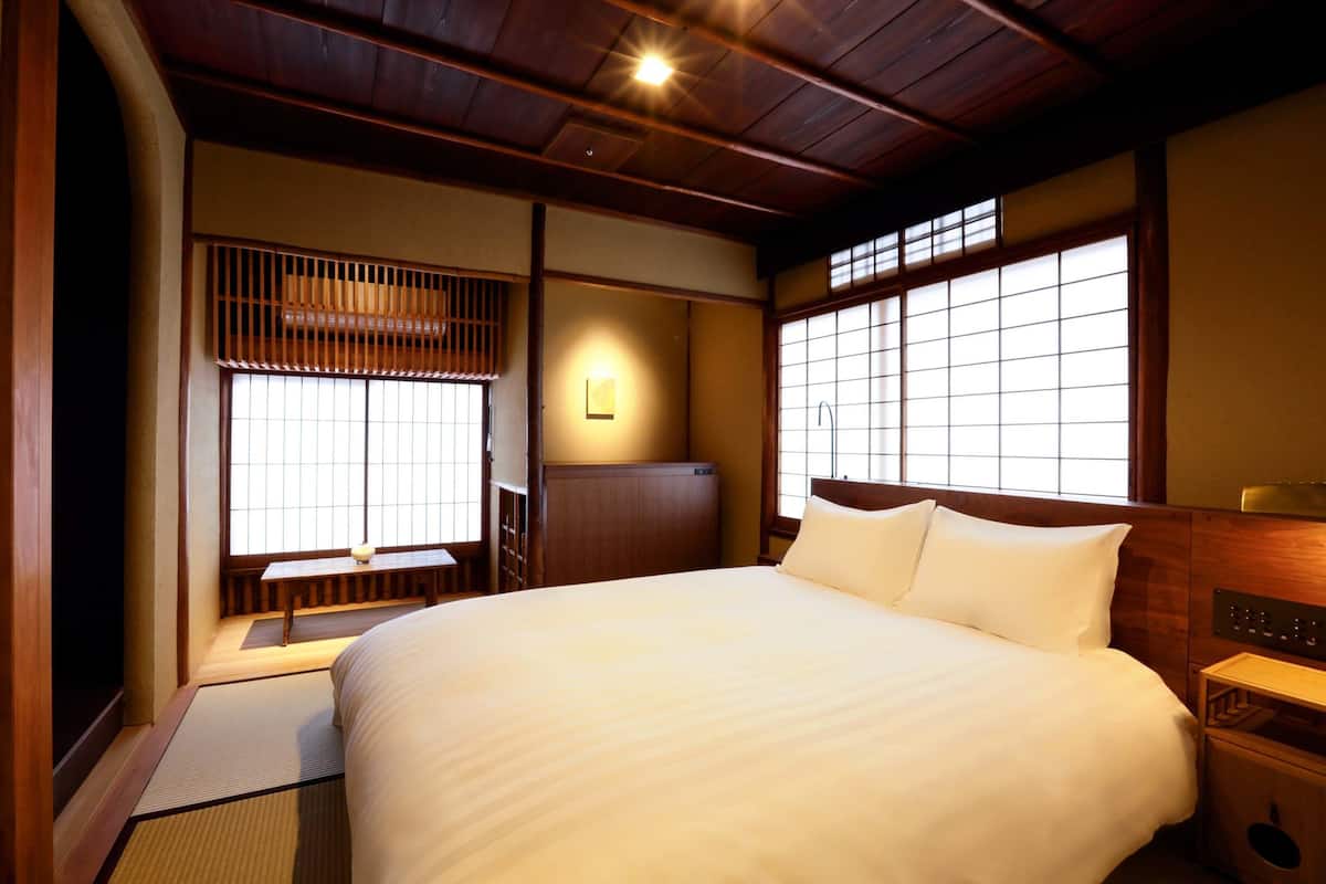 Een traditionele Japanse slaapkamer met tatami vloeren, een groot bed met wit beddengoed, schuifdeuren en een houten hoofdbord.