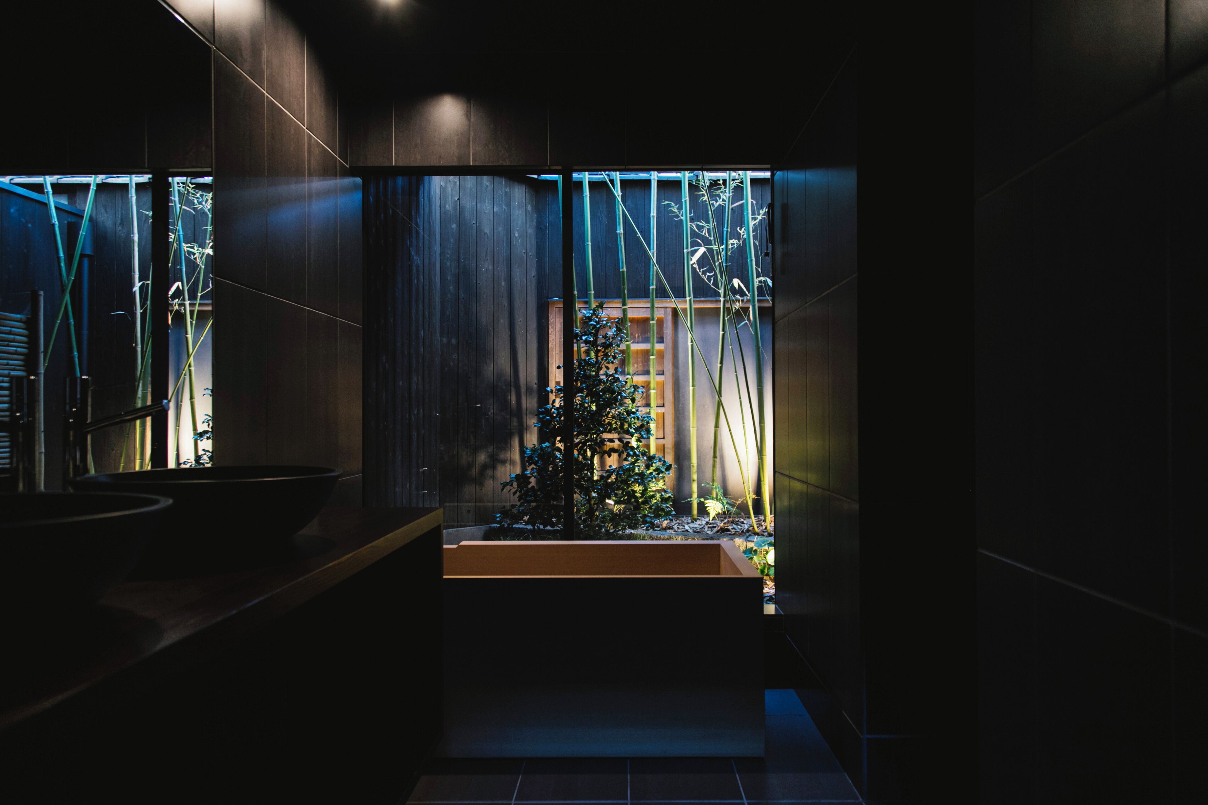 A modern bathroom with a dark interior, a wooden bench, and a view of bamboo outside.