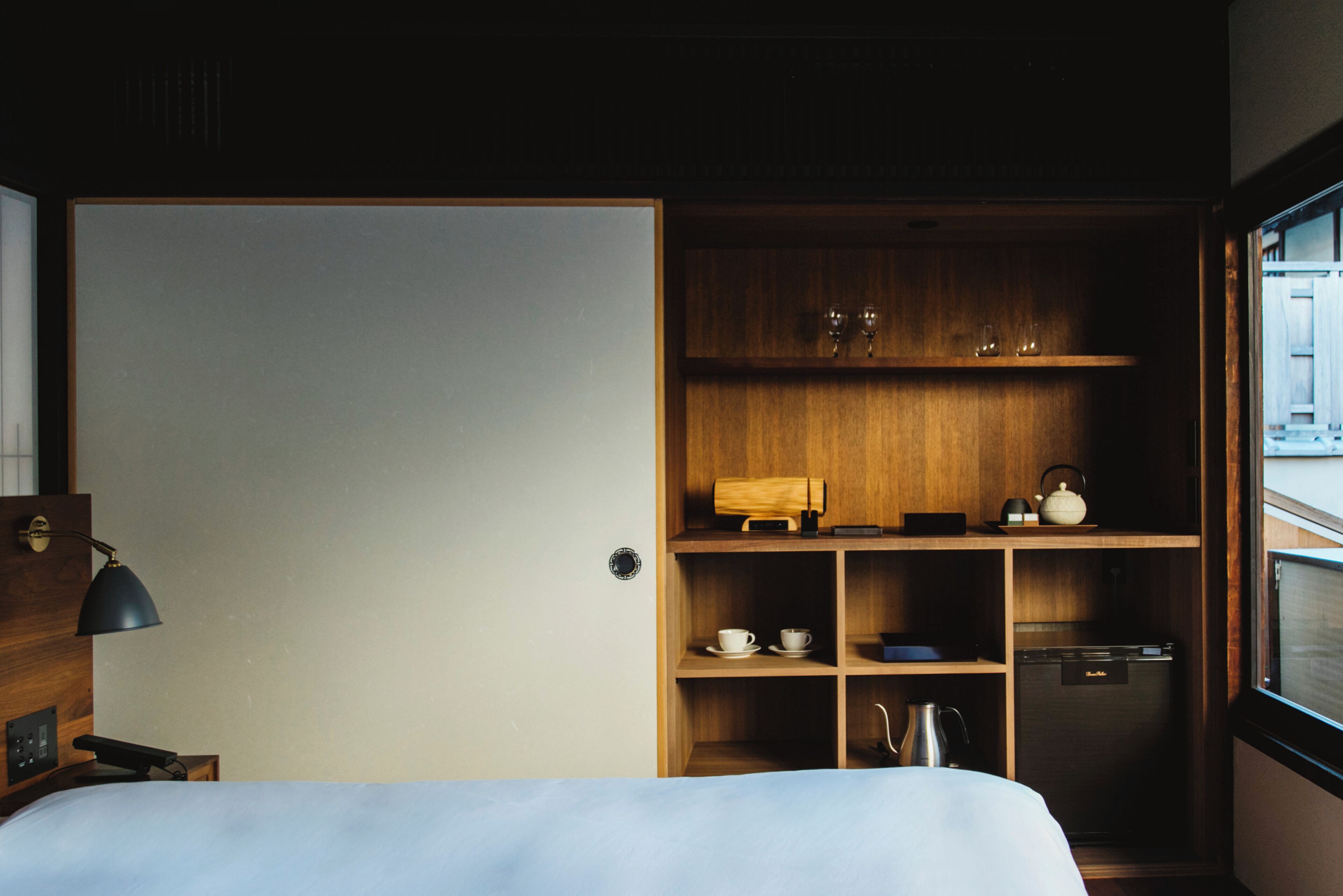 A hotel room with a bed, a desk with a lamp, a wardrobe, and a kitchenette area with a coffee maker and a kettle.