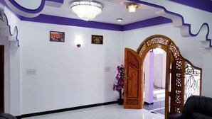 A room with a white floor, purple accents on the ceiling, a chandelier, a wooden door, and a black sofa. - SNP Star Guesthouse (Trincomalee)
