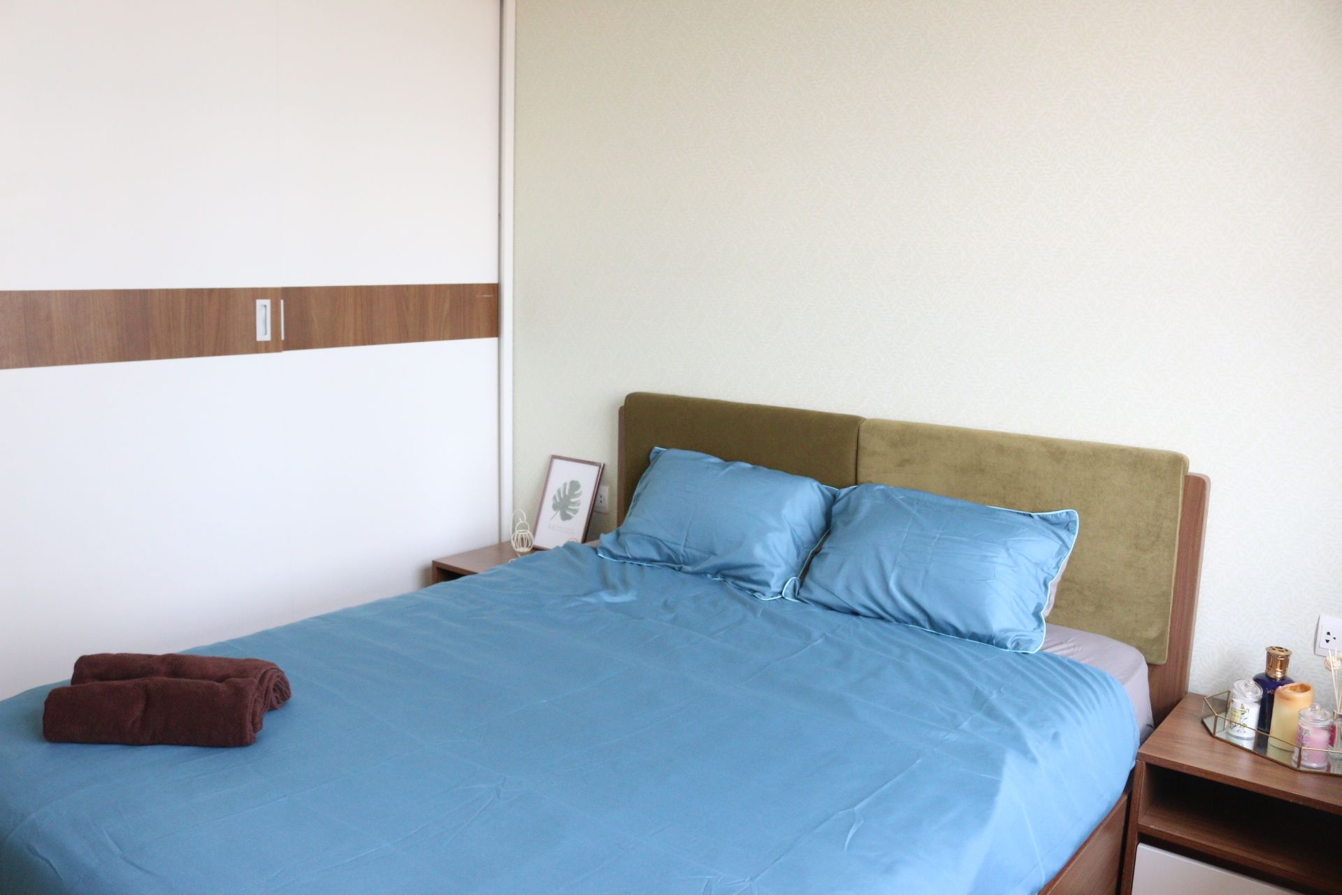 A neatly made bed with a blue bedspread and two pillows, a brown towel, and a bedside table with a lamp and a framed picture.