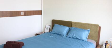 A neatly made bed with a blue bedspread and two pillows, a brown towel, and a bedside table with a lamp and a framed picture.