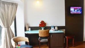 A room with a dining table, chairs, a small cabinet, a TV mounted on the wall, and a window with curtains. - Ashraya International Hotel (Bengaluru)