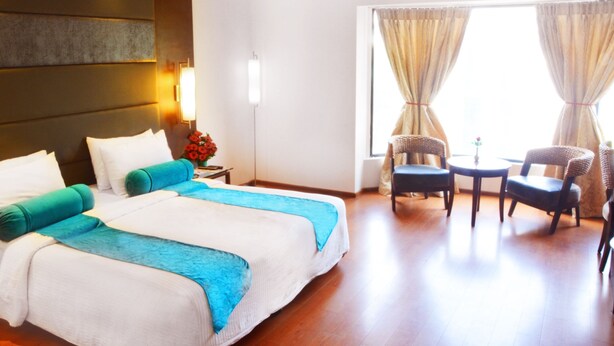 A hotel room with a large bed, two chairs, a small table, a nightstand, and a window with curtains. - Ashraya International Hotel (Bengaluru)