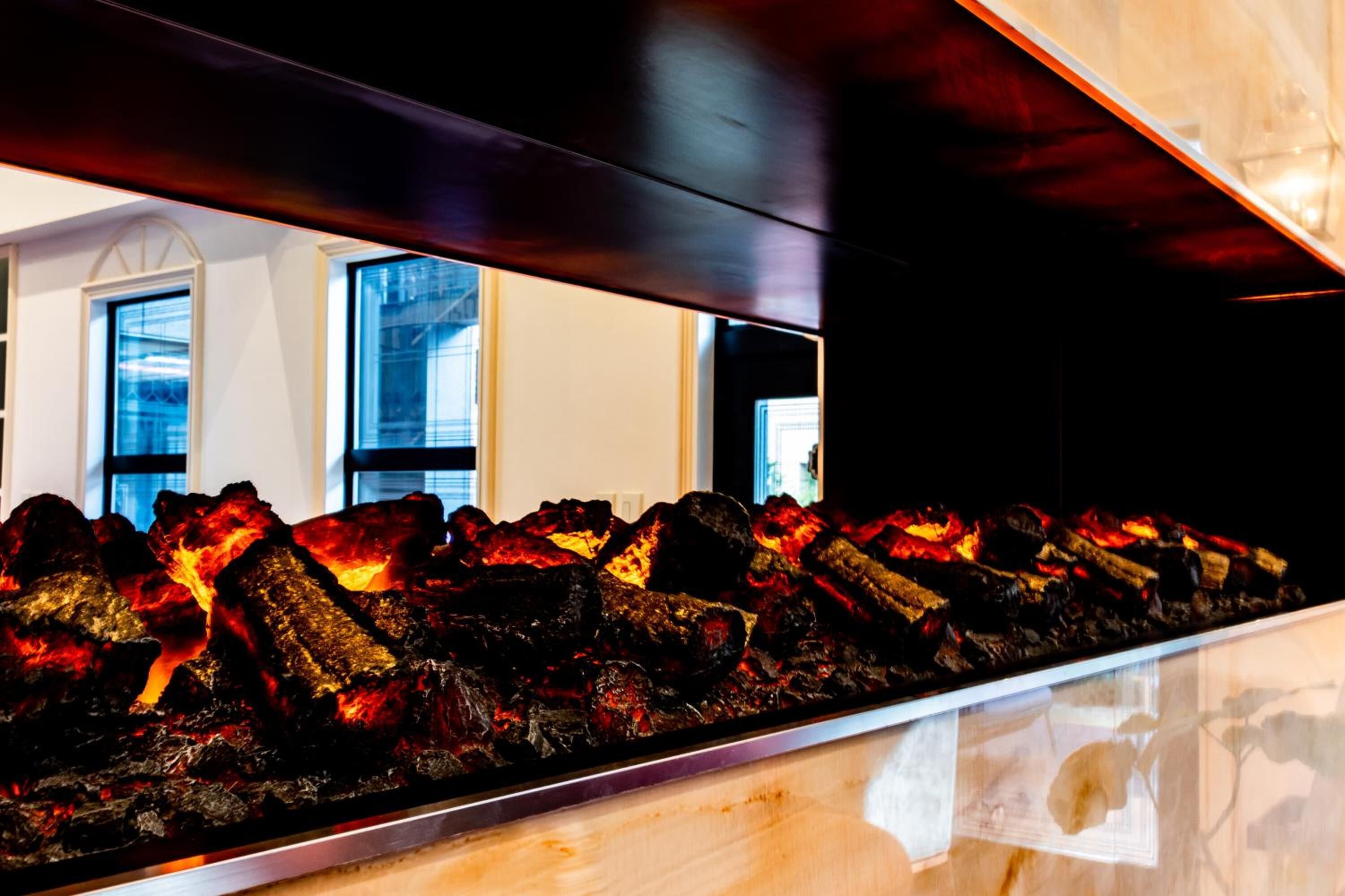 A charcoal grill with visible flames, set against a modern interior with large windows and a wooden ceiling.