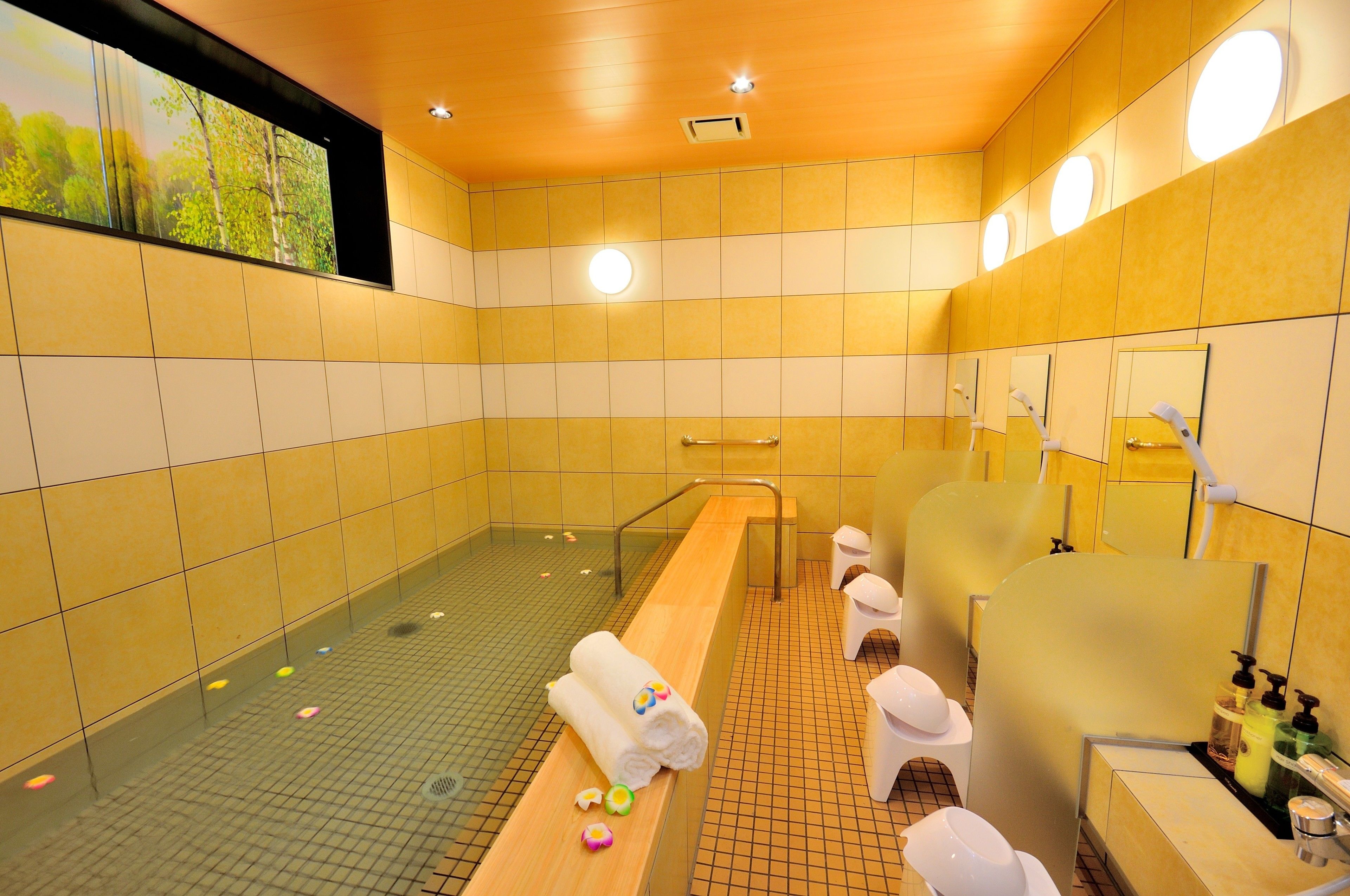 A modern bathroom with yellow tiled walls, a large mirror, and a long bench with towels.