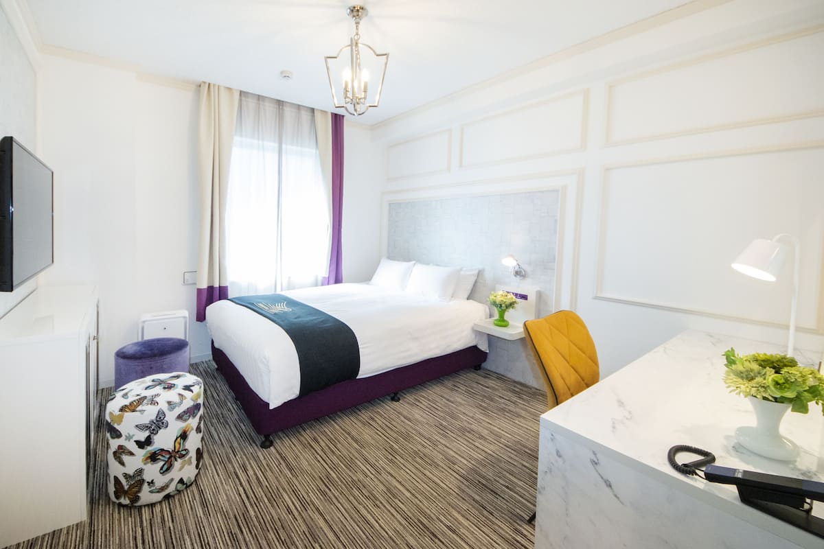 Habitación de hotel con cama, escritorio, silla, televisor y ventana con cortinas.