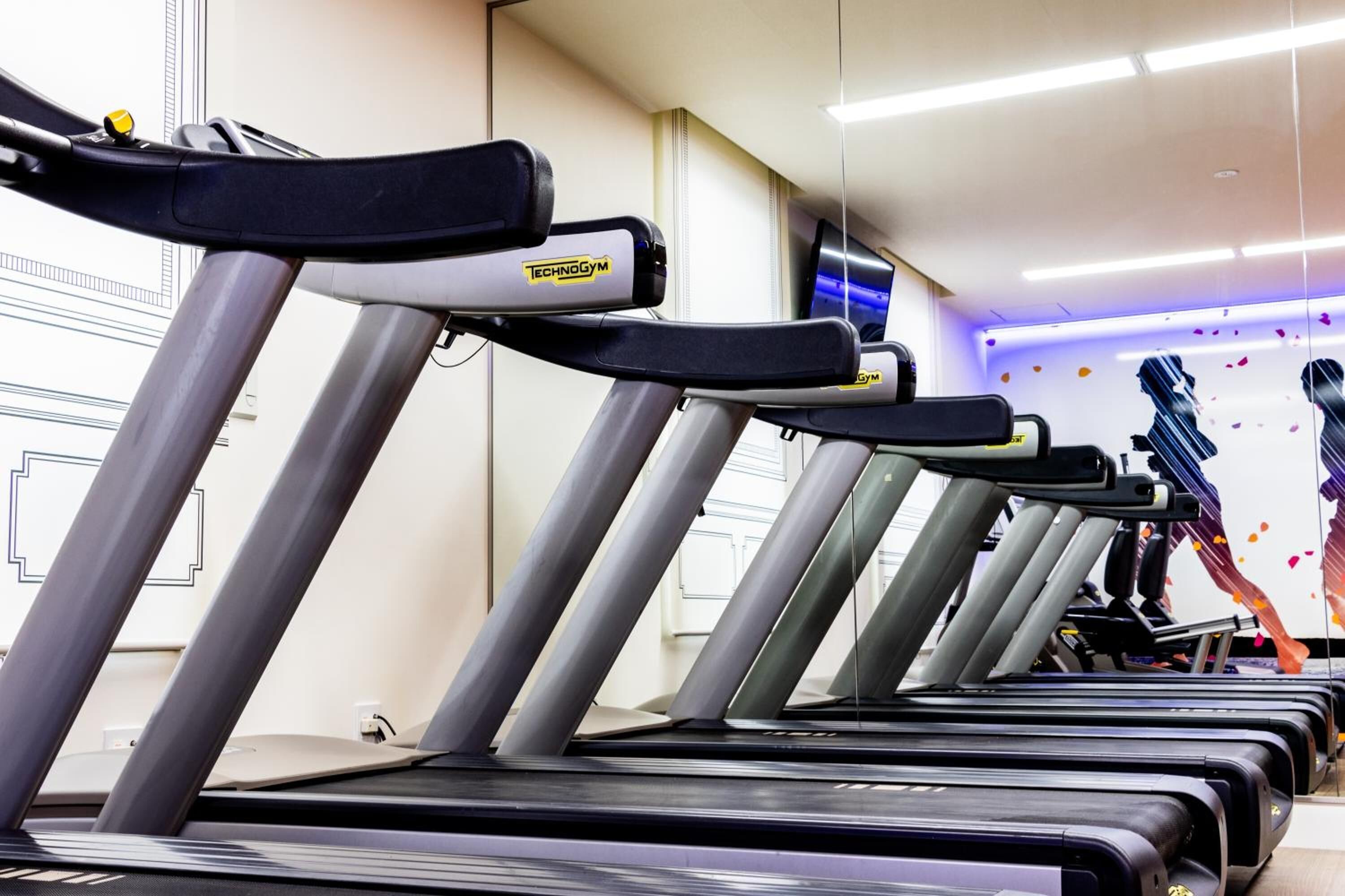 Varias cintas de correr en un gimnasio con un espejo grande y una pared decorativa con siluetas.