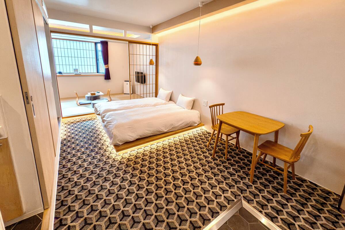Une chambre moderne avec un grand lit, une petite table et une chaise. Il y a une fenêtre avec des rideaux et une vue sur l’extérieur.