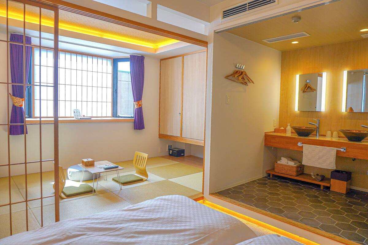 Une chambre d’hôtel avec un sol recouvert de tatamis, un lit, une petite table avec une chaise, une fenêtre avec des rideaux violets et une salle de bain avec un meuble-lavabo en bois et un grand miroir.