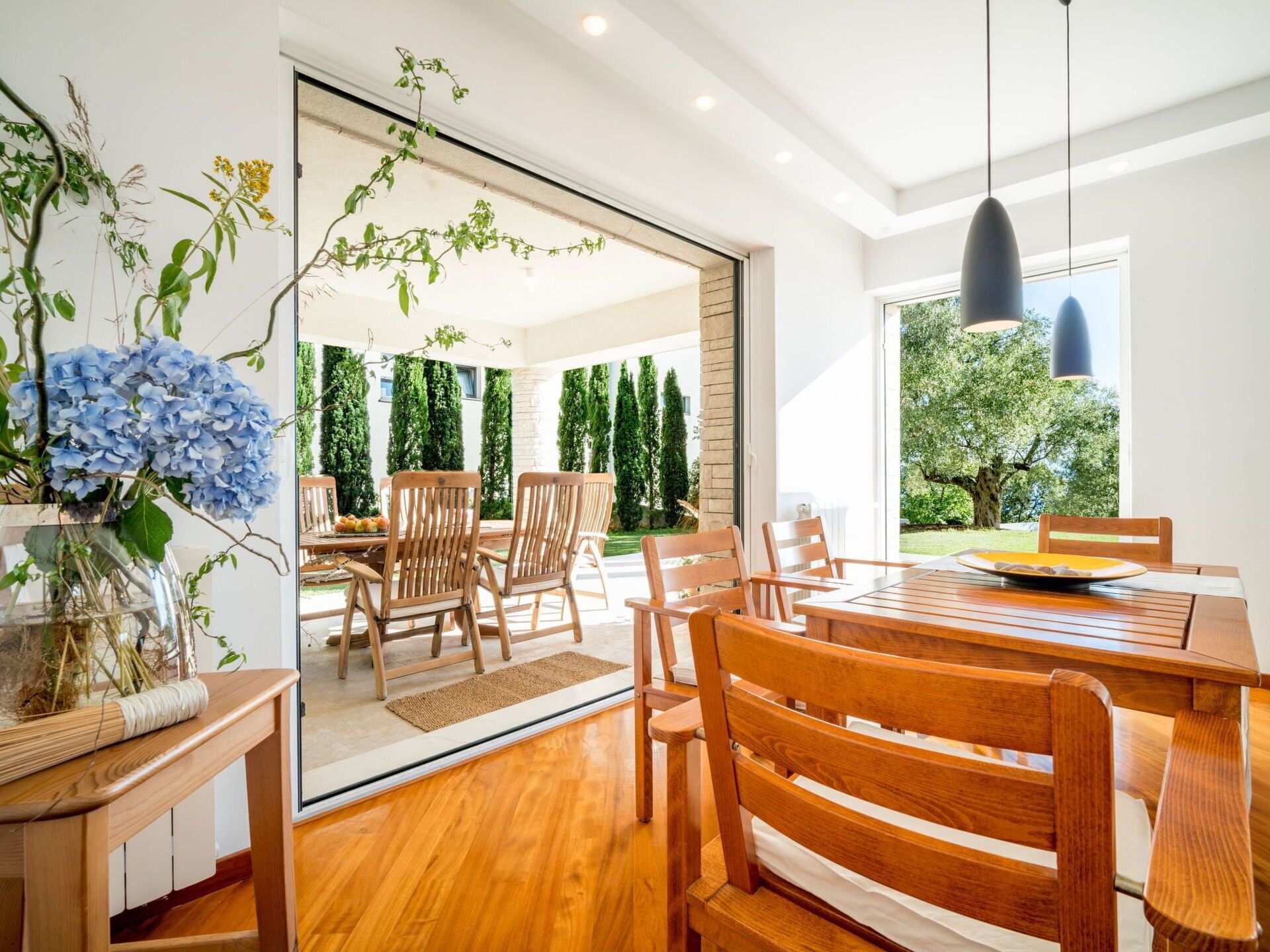 A modern dining area with wooden furniture and a glass door leading to an outdoor space with greenery.