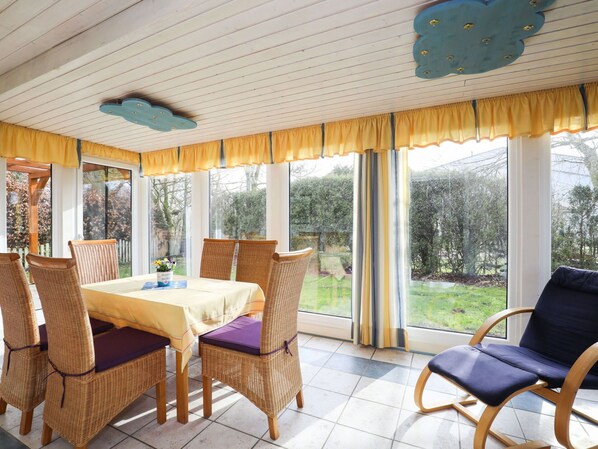Ein Essbereich mit Korbstühlen und einem Tisch, großen Fenstern mit Vorhängen und Blick auf den Garten.