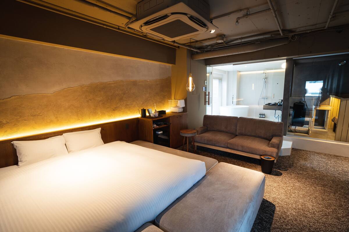A hotel room with a large bed, a sofa, a small table, and a bathroom visible through an open door.