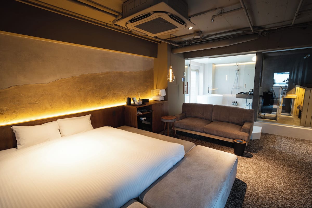 A hotel room with a large bed, a sofa, a small table, and a bathroom visible through a glass door.