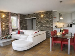 Una sala de estar moderna con un sofá blanco, sillas rojas, una mesa de comedor de vidrio y una zona de cocina con armarios blancos y una pared de ladrillo.