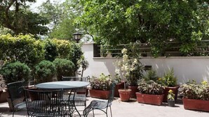 A patio area with a round table and chairs, surrounded by potted plants and a white wall. - Perch Arbor Suites (Gurugram)