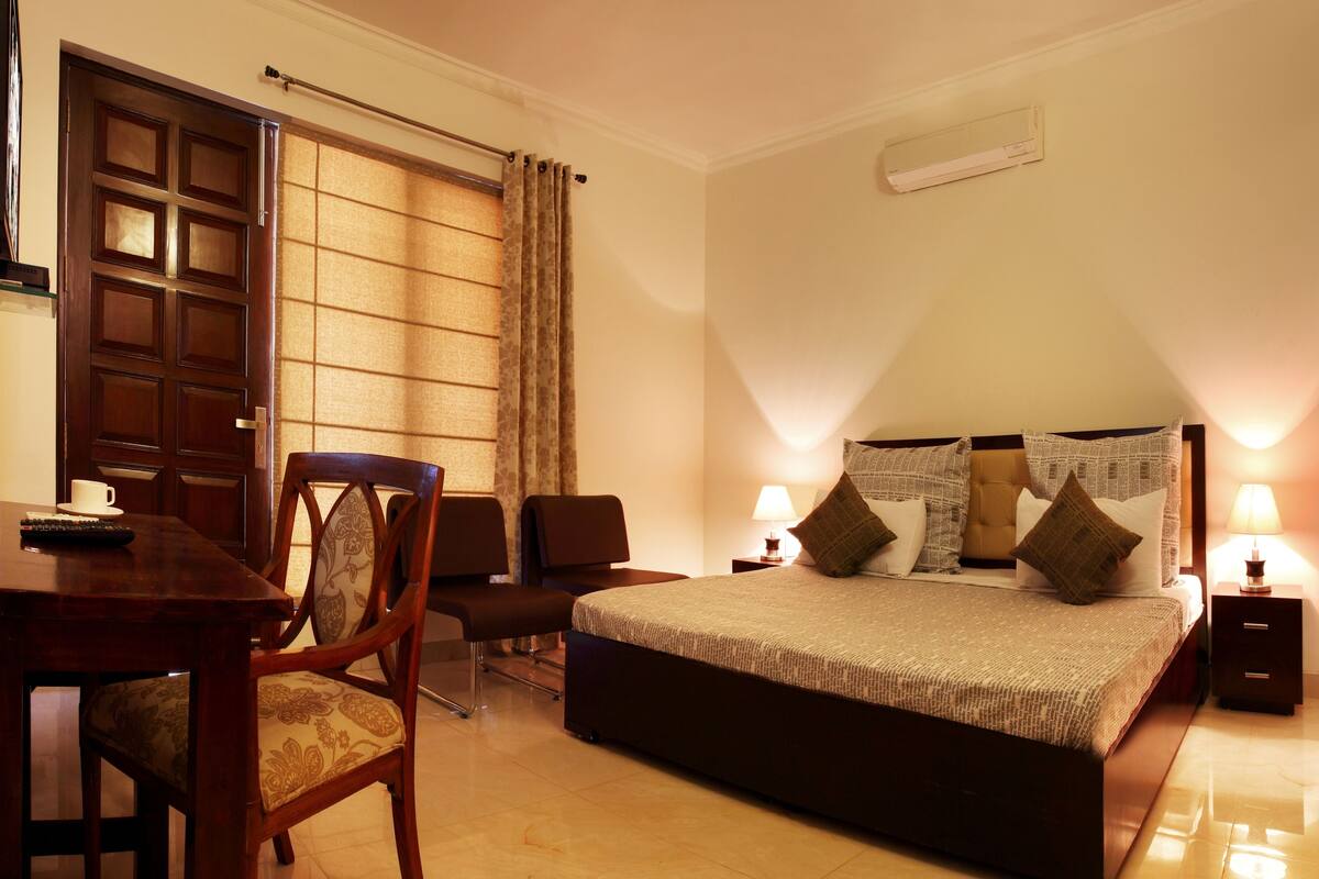 A hotel room with a large bed, a desk with a chair, a window with curtains, and a bedside table with a lamp.