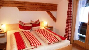 Une chambre avec un plafond en bois, un lit recouvert d’une couette à carreaux rouges et blancs, une table de chevet avec une lampe, et une fenêtre ornée de rideaux rouges et blancs.
