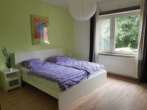 Quarto com cama, mesa de cabeceira, planta em vaso, janela com cortinas e luminária pendente.